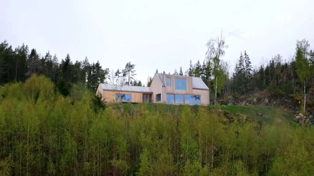 Stiligt - Ett modernt trähus med stora fönster ligger på en kulle omgiven av gröna träd och skog under en mulen himmel.