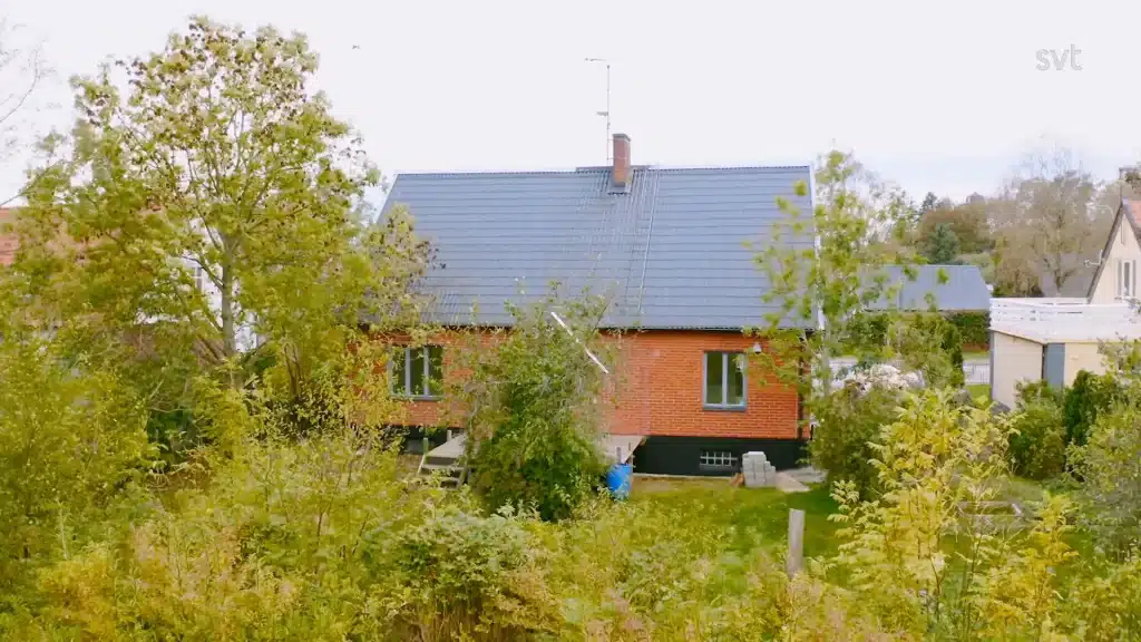 Stiligt - Ett rött tegelhus med mörkgrått tak är omgivet av gröna träd och buskar en molnig dag.