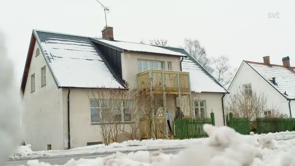 Stiligt - Ett tvåvåningshus med snötak, träbalkong och grönt staket, omgivet av snötäckt mark, en molnig vinterdag.