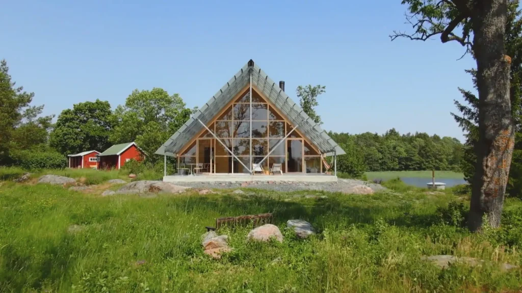 Stiligt - A-ramhus med stora glasfönster som ligger i ett gräsbevuxet fält, omgivet av träd, med en liten röd byggnad i närheten under en klarblå himmel.