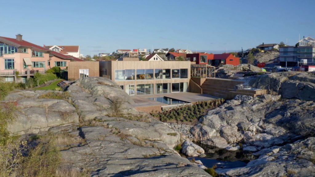 Stiligt - Ett modernt tvåvåningshus med stora glasfönster står bland stenig terräng, omgivet av andra hem och klarblå himmel.