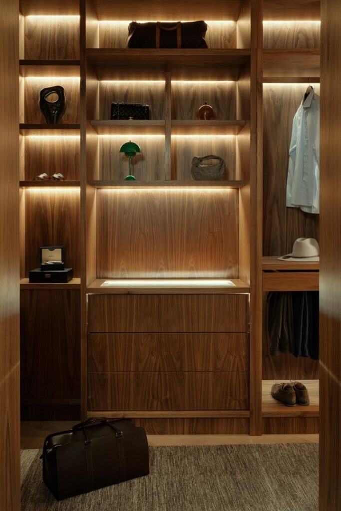 Stiligt - En modern klädkammare i trä i sekelskiftesstil med upplysta hyllor, lådor, hängande kläder, hatt, skor, väskor och prydnadsföremål, med en mörk läderväska på golvet.