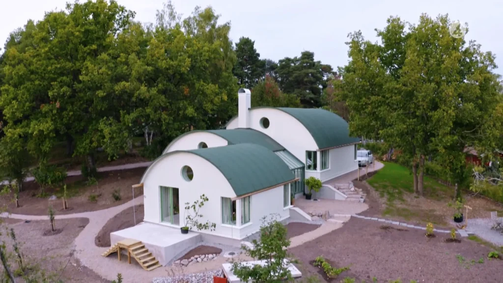 Stiligt - Två moderna vita hus med svängda gröna tak, runda fönster och flera ingångar, omgivna av träd och landskap i en lantlig miljö.