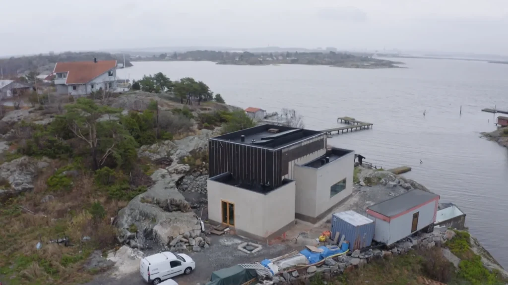 Stiligt - Ett modernt hus under uppbyggnad sitter på en stenig strandlinje med utsikt över vattnet; byggmaterial och en vit skåpbil syns bredvid byggnaden.