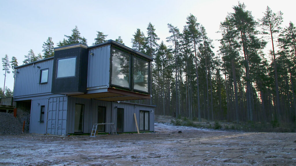Stiligt - Ett modernt hus i två våningar av fraktcontainrar ligger på en glänta nära en skog, med stora fönster och en ofullbordad exteriör.