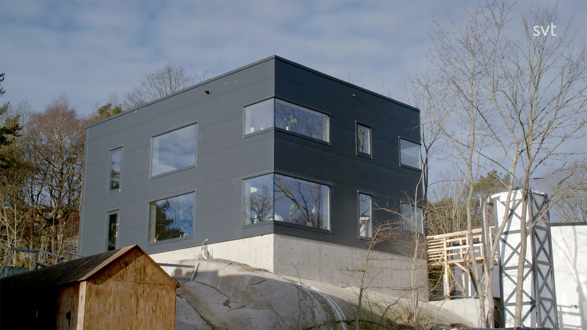 Stiligt - Ett modernt, mörkgrått, lådformat hus med stora fönster ligger på en stenig sluttning bredvid bladlösa träd under en delvis molnig himmel.