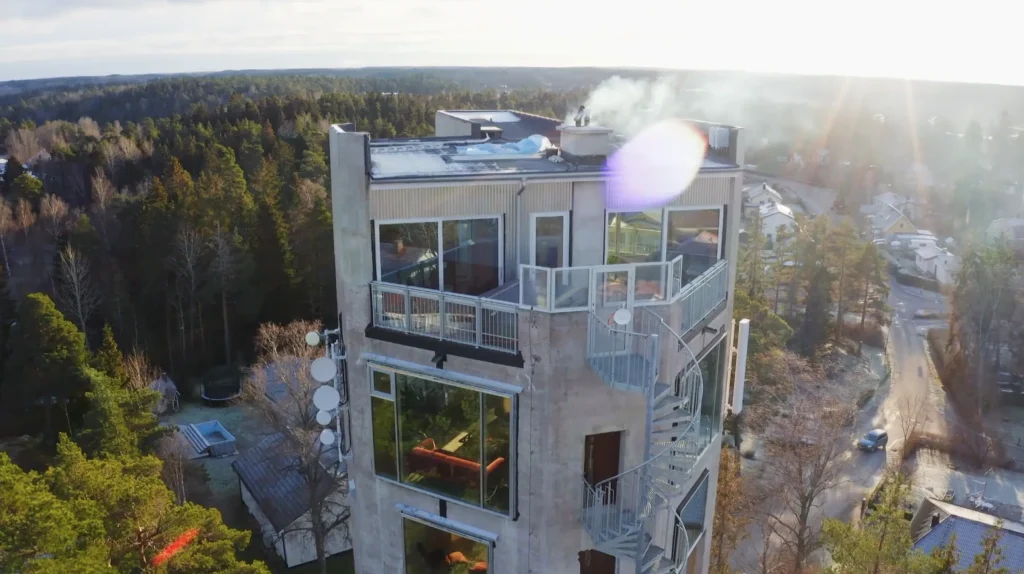 Stiligt - En modern flervåningsbyggnad med stora fönster och en yttre spiraltrappa är omgiven av träd; rök stiger upp från taket.