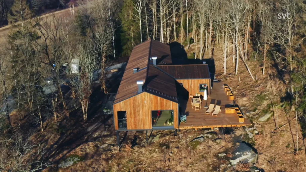 Stiligt - Ett modernt trähus med ett stort däck ligger i ett skogsområde med lövlösa träd och utemöbler synliga på däck.