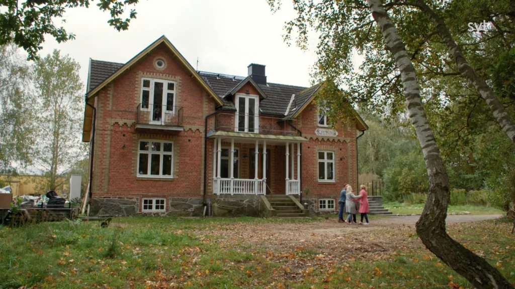 Stiligt - Ett stort rött tegelhus med vita lister står på en gräsbevuxen gård när fyra personer går tillsammans på stigen framför det.