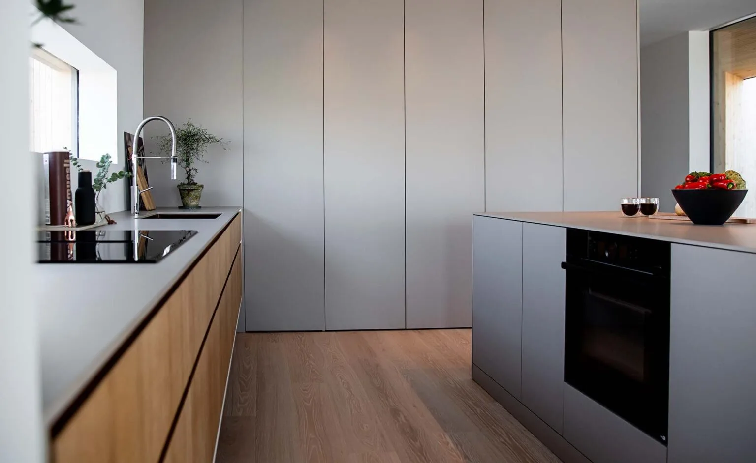 Stiligt - Modernt kök med minimalistiska skåp, inbyggd ugn, ö, induktionshäll, trägolv och en skål med råvaror på bänken.