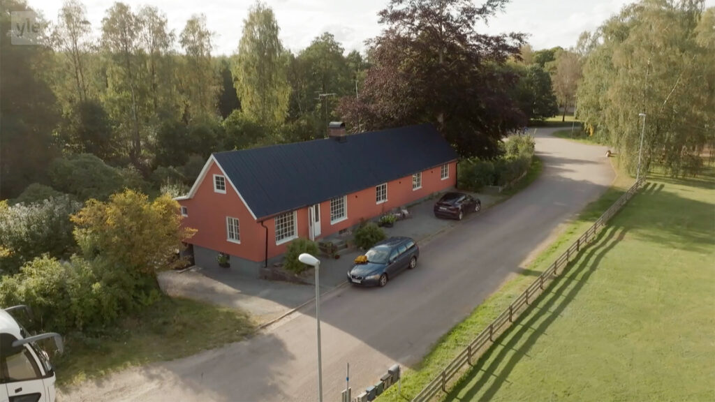 Stiligt - Ett rött hus med mörkt tak ligger bredvid en väg, omgivet av träd. Två bilar står parkerade utanför, och ett trästaket löper längs fastigheten.