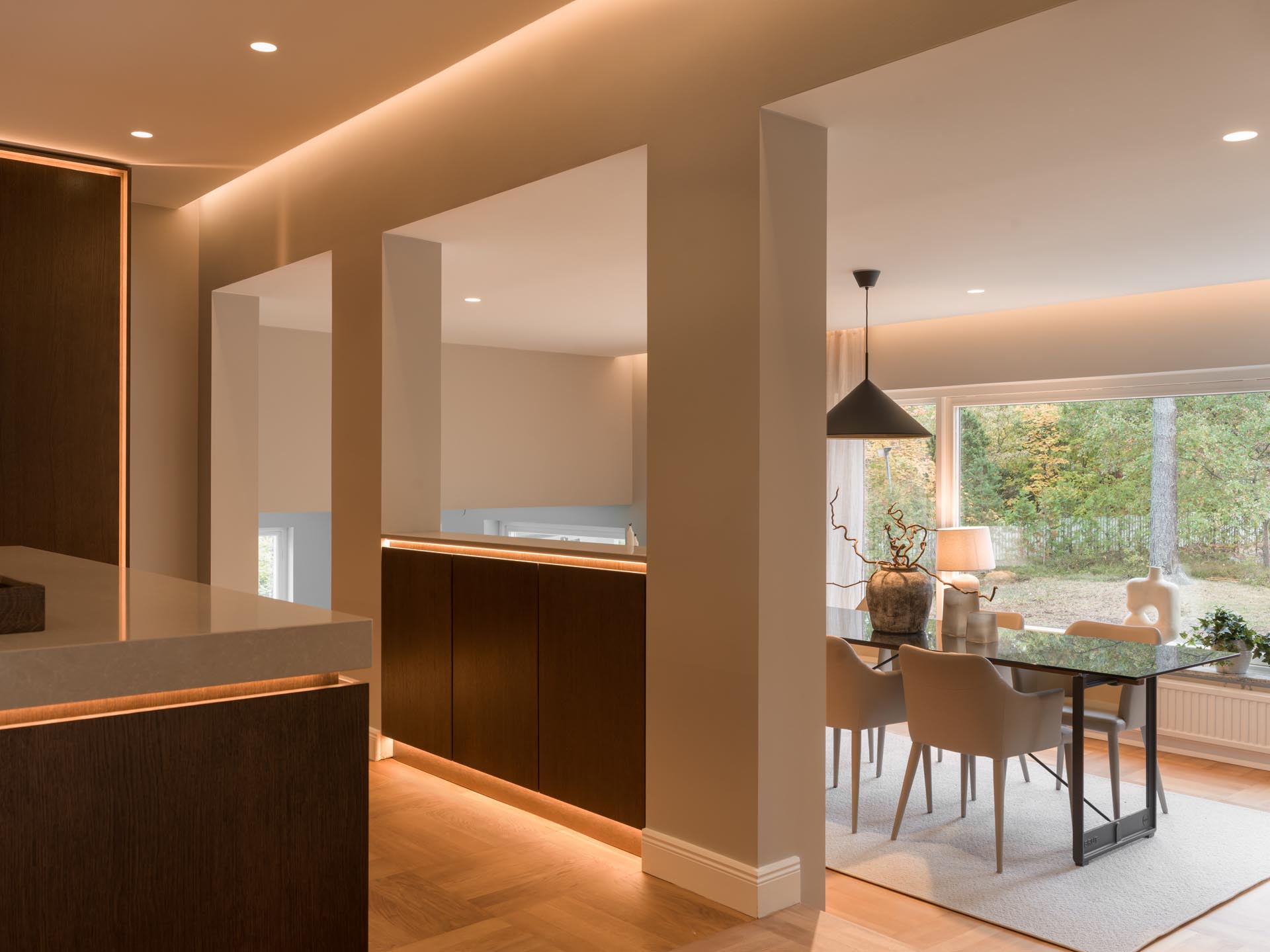 Stiligt - Modern inredning i öppen planlösning med varm belysning, mörka träskåp och en matplats med ett glasbord och beige stolar. Träkök med pocketdörrar öppnar upp mot stora fönster med utsikt över träden utanför.