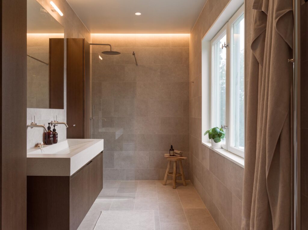 Stiligt - Modernt badrum med stort beige kakel, duschkabin, dubbla handfat, träpall och fönster. Handdukar och en liten krukväxt sitter på fönsterbrädan och kompletterar den intilliggande träkök med pocketdörrar.