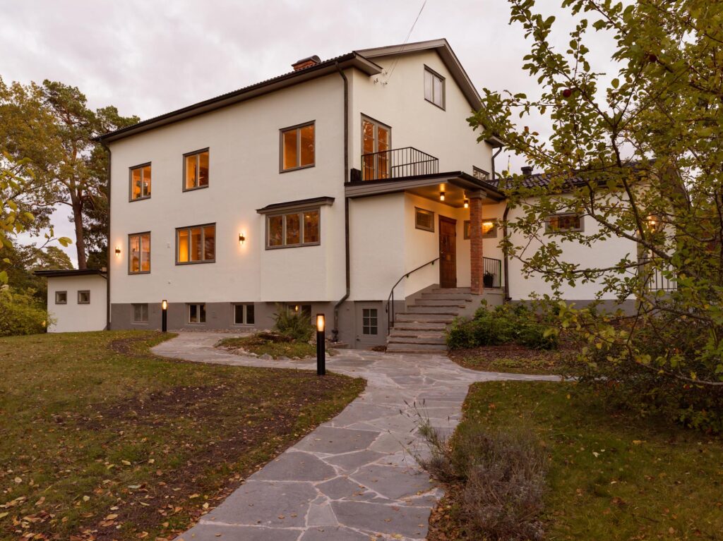 Stiligt - Ett tvåvånings krämfärgat hus med stora fönster, en veranda och en stengång med en träkök med pocketdörrar inuti, omgiven av gräs, träd och utomhusbelysning i skymningen.