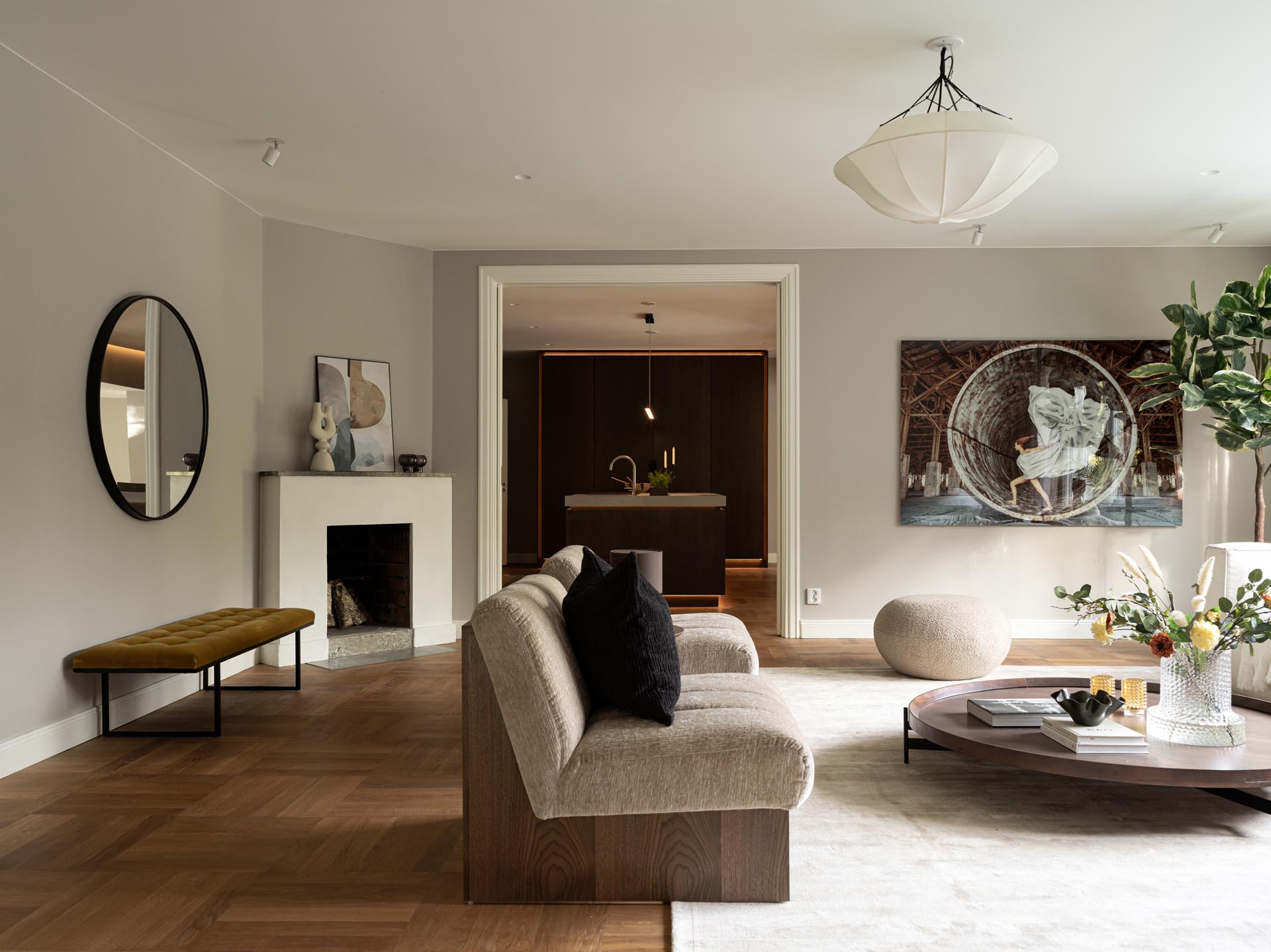 Stiligt - Modernt vardagsrum med neutrala toner, beige soffa, runt soffbord, väggkonst, öppen spis, samt utsikt mot ett intilliggande träkök med pocketdörrar; minimal och modern inredning.