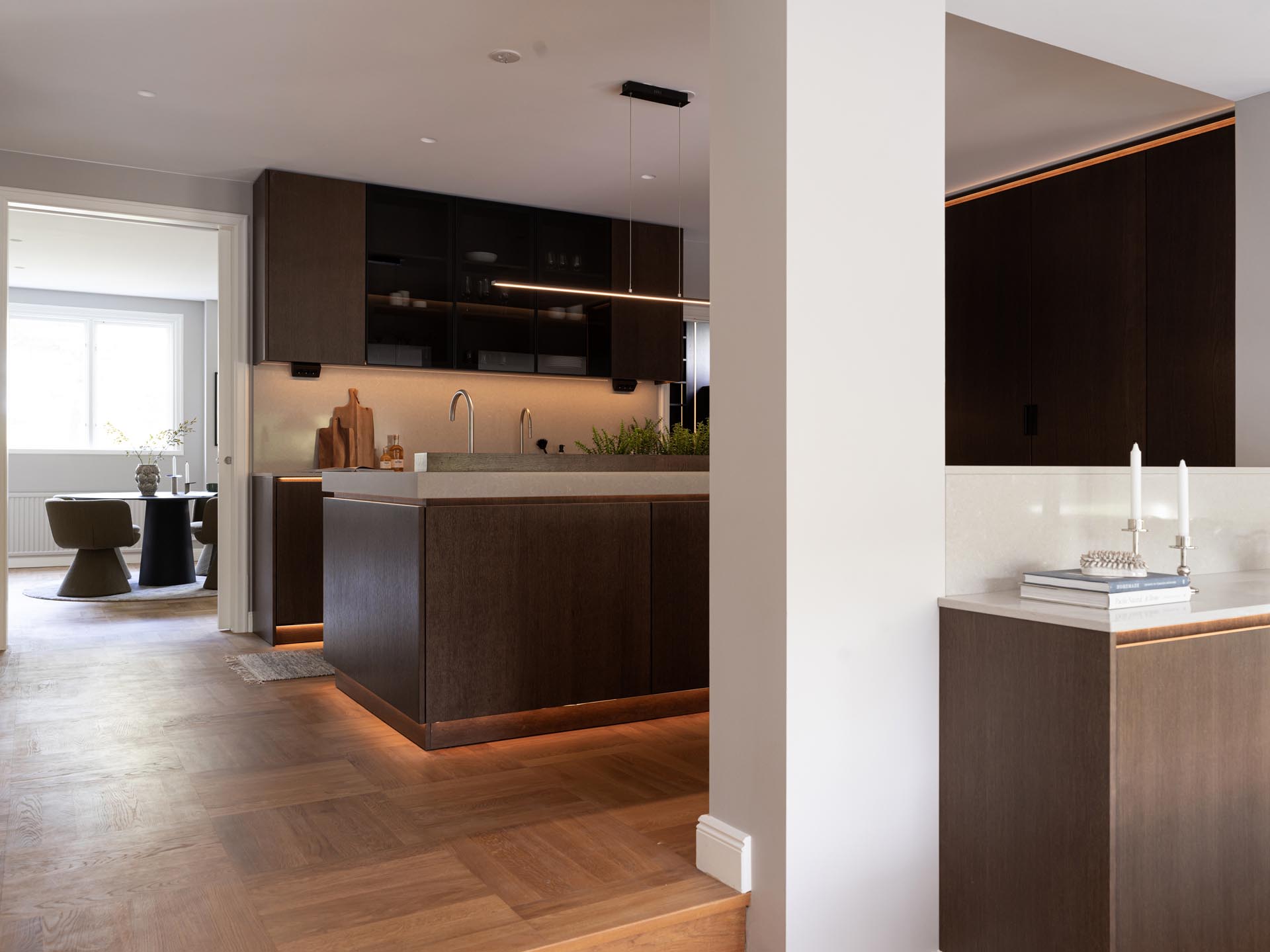 Stiligt - Modernt kök med skåp i mörkt trä, en stor ö och belysning under skåpet, med minimal inredning och en intilliggande matplats – denna träkök med pocketdörrar erbjuder både stil och smarta utrymmesbesparande lösningar.