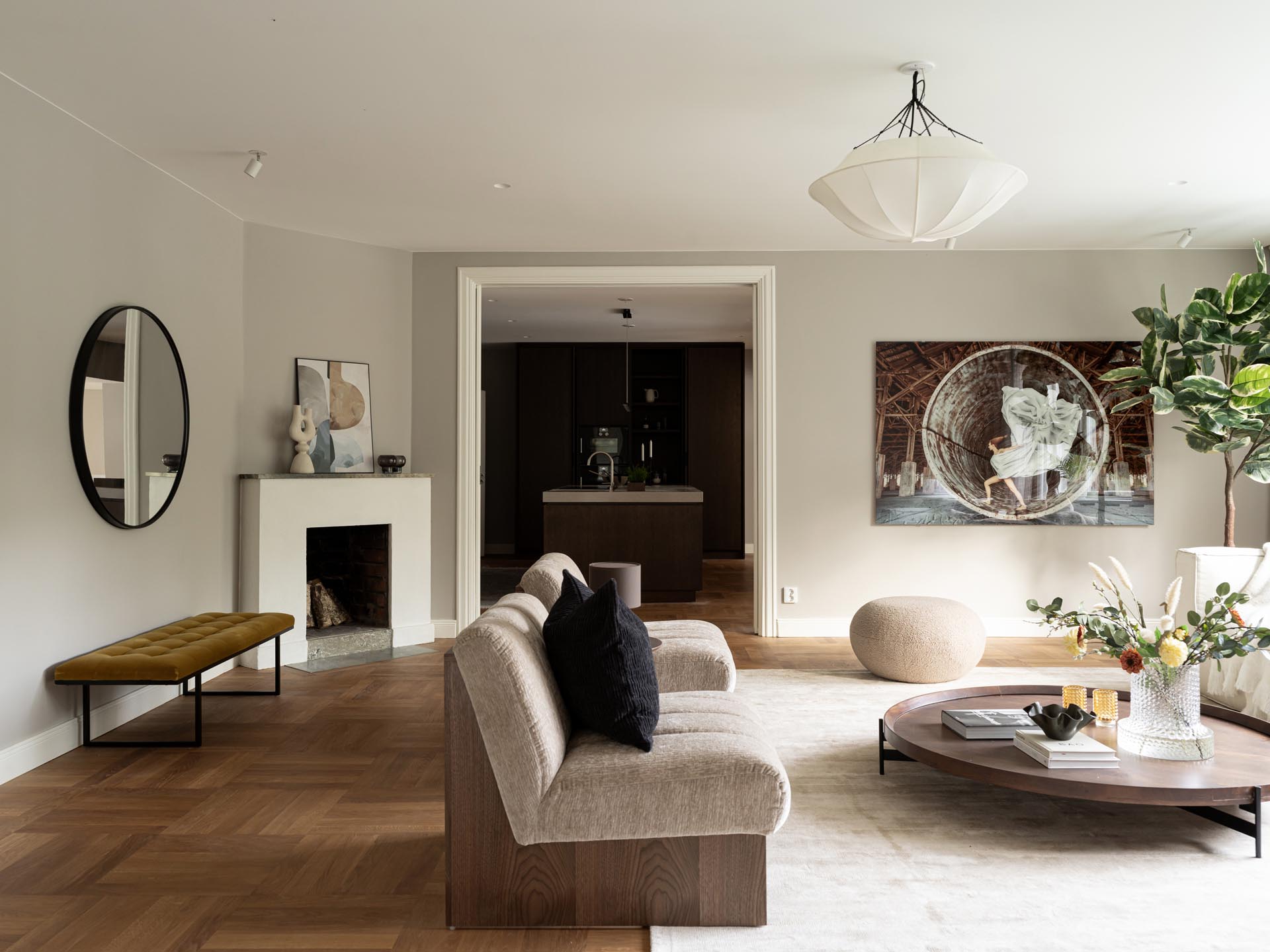 Stiligt - Modernt vardagsrum med neutrala toner, en beige soffa, runt soffbord, väggkonst, öppen spis, stor planta, och en öppen utsikt mot en träkök med pocketdörrar.