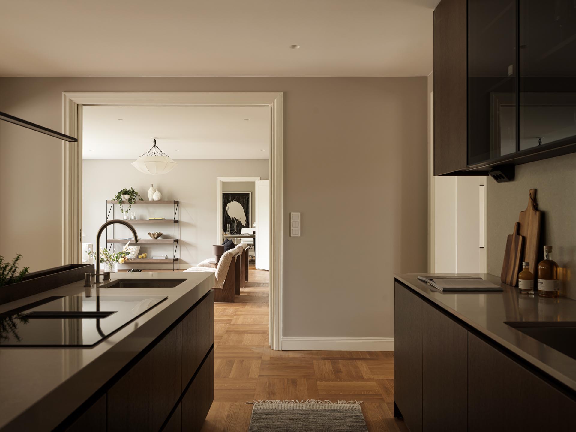 Stiligt - Modernt kök med mörka skåp och bänkskivor, tittar in i ett vardagsrum med trägolv och minimalistisk inredning. Naturligt ljus kommer in från fönstren i det intilliggande rummet, vilket framhäver träkök med pocketdörrar för sömlös stil.