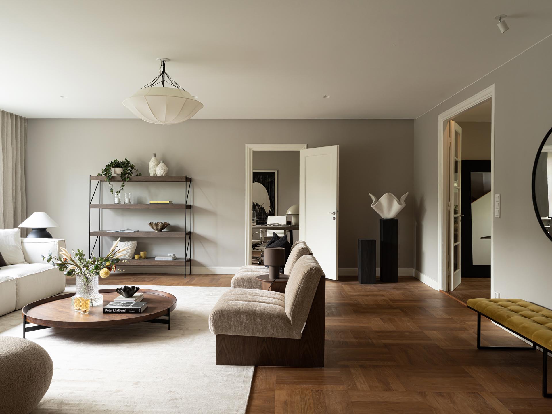 Stiligt - Modernt vardagsrum med neutrala toner, med en beige soffa, två stolar, ett runt soffbord, en metallhylla och minimal inredning på trägolv.