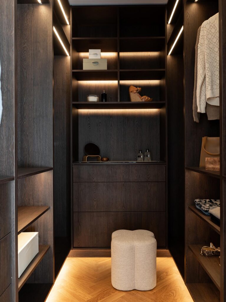 Stiligt - En modern klädkammare med hyllor och lådor i mörkt trä, upplyst av inbyggd belysning, har en liten beige stoppad pall på ett trägolv – perfekt komplement till alla träkök med pocketdörrar för sömlös elegans.