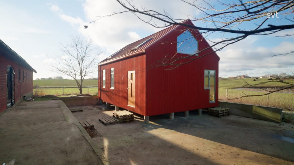 Stiligt - Ett litet rött hus med sluttande tak står på betongblock på en öppen yta intill en större byggnad, omgiven av åkrar och kala träd.