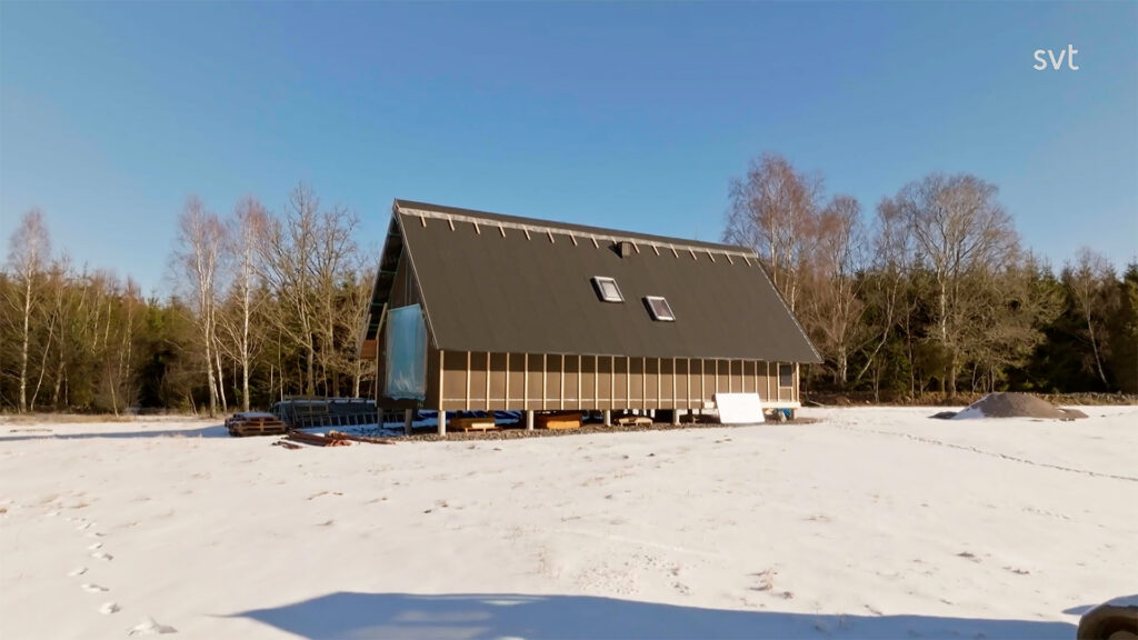 Stiligt - Ett modernt hus i A-ram med mörk takbeläggning står på en snöig åker, omgiven av bladlösa träd och högar av material i närheten under en klarblå himmel.