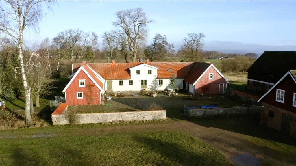 Stiligt - Flygfoto över ett lantligt huskomplex med röda och vita byggnader, omgiven av gräs, träd och en stenmur under en klar himmel.