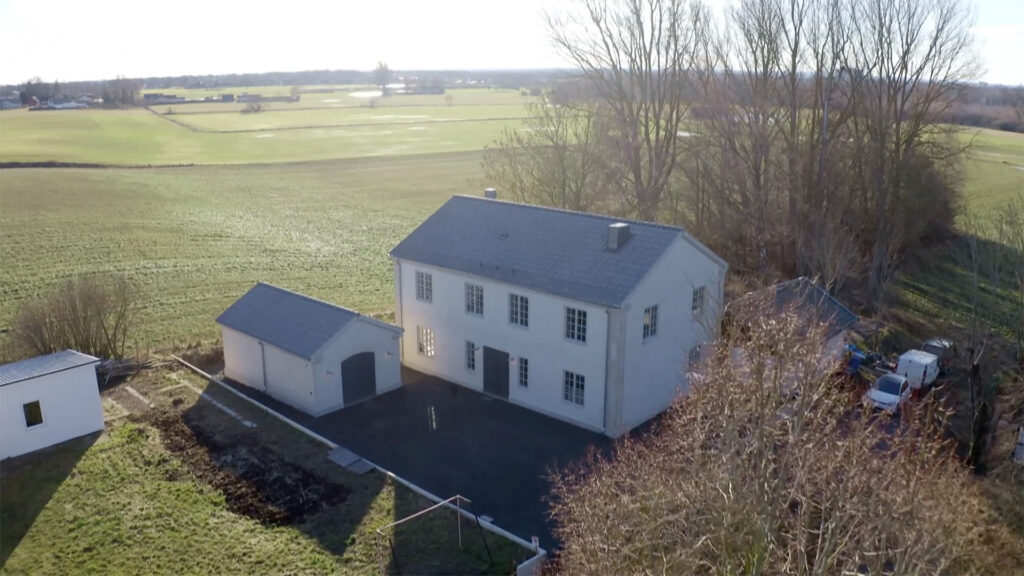 Stiligt - Flygfoto över ett vitt tvåvåningshus med ett fristående garage, beläget intill öppna fält och kantat av lövlösa träd.