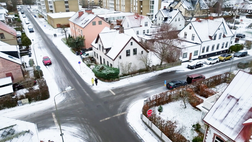 Stiligt - Flygfoto över ett snöigt bostadsområde med flera hus, bilar parkerade längs gatorna och fotgängare som korsar en korsning.