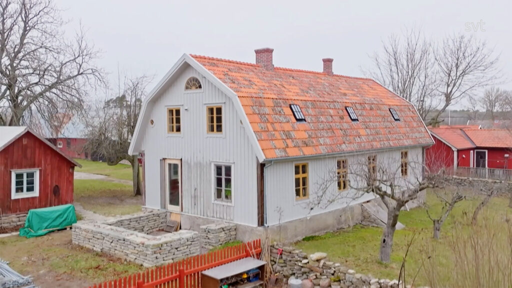 Stiligt - Ett vitt tvåvåningshus i ladugårdstil med ett rött tegeltak, omgivet av en gård, träd och ett rött trästaket.