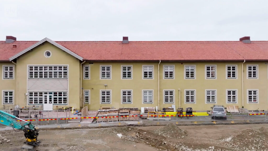 Stiligt - En tvåvåningsbyggnad med rött tegeltak är under uppbyggnad, med byggutrustning och material framför och smutsmark i förgrunden.