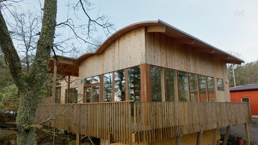 Stiligt - Ett modernt trähus med stora fönster och ett böjt tak, omgivet av träd och med ett inhägnat däck.