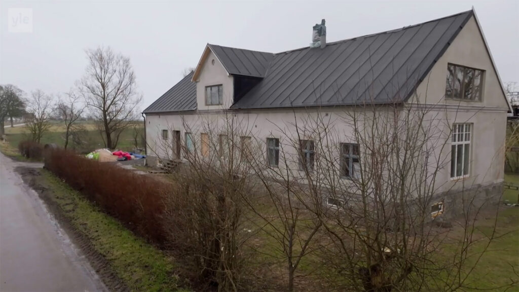 Stiligt - Ett stort, ljust hus med mörkt plåttak står bredvid en landsväg, kantad av lövlösa buskar och kala träd en molnig dag.