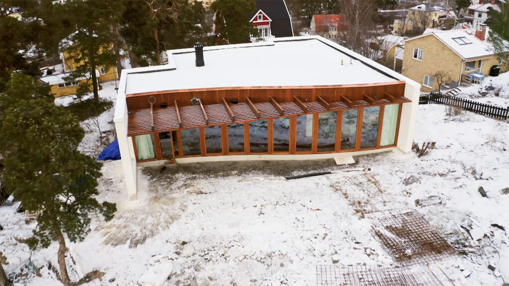Stiligt - Ett modernt, rektangulärt hus med platt tak och stora glasfönster står på en snörik gård omgiven av träd och grannhus.