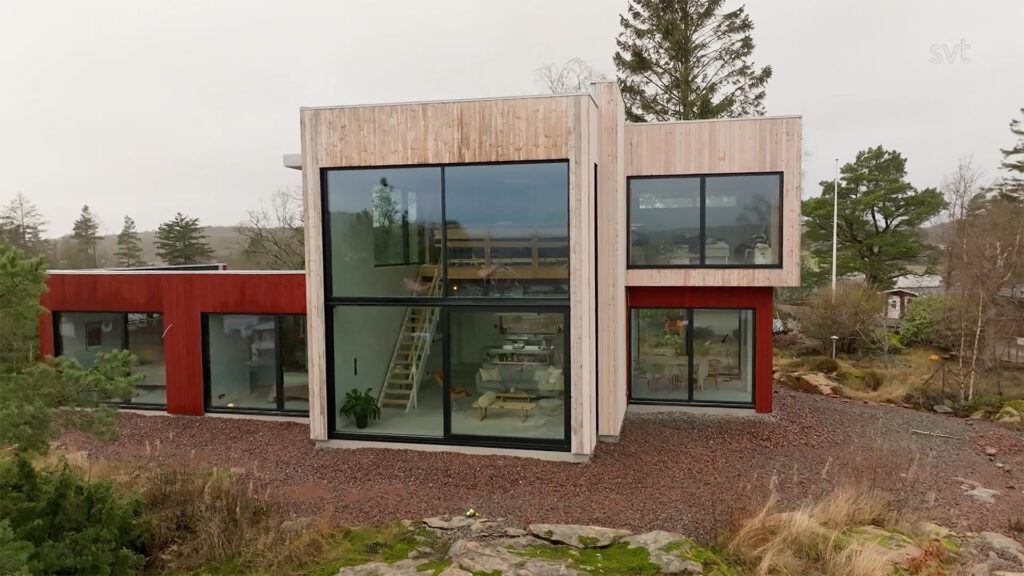 Stiligt - Modernt tvåvåningshus med stora glasfönster och träpanel, omgivet av träd och stenig terräng under en mulen himmel.