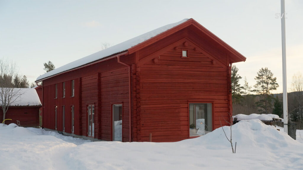 Stiligt - Ett stort rött trähus med snötäckt tak och omgivningar, i ett lantligt vinterlandskap med träd i bakgrunden.