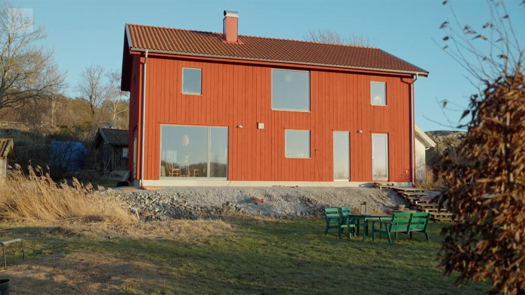Stiligt - Ett tvåvånings rött hus med sluttande tak och stora fyrkantiga fönster står på en stenig tomt med gröna stolar och ett bord på gräset framför.