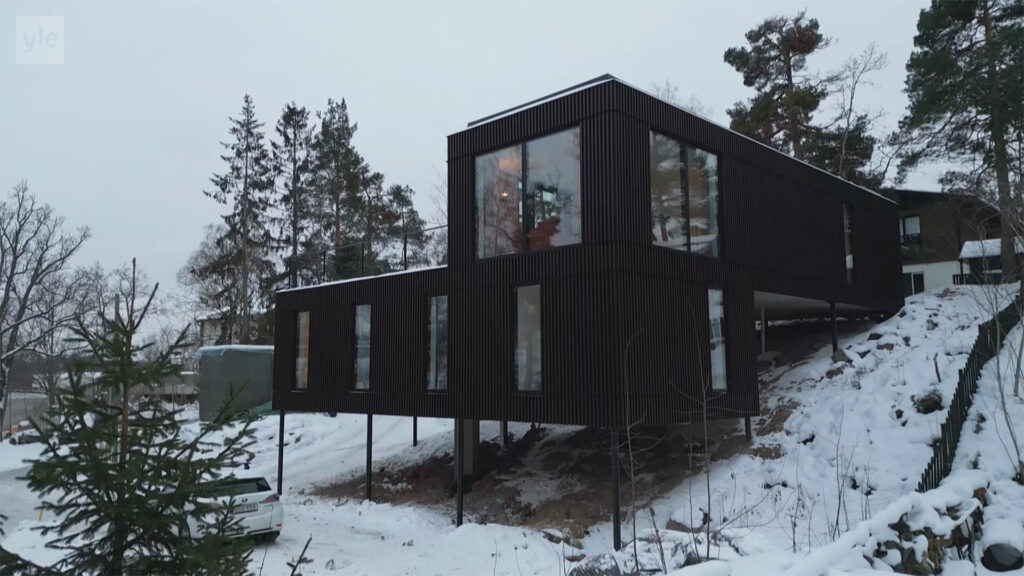 Stiligt - Ett modernt svart hus på pålar står i ett snöigt, skogslandskap, med stora fönster och minimalistisk arkitektur.