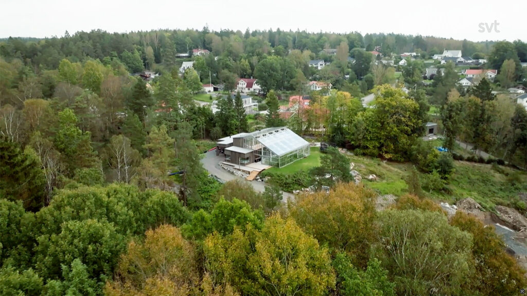 Stiligt - Ett modernt hus med ett stort glasväxthus är omgivet av träd och grönska, beläget i ett förortsområde med andra hus synliga i bakgrunden.