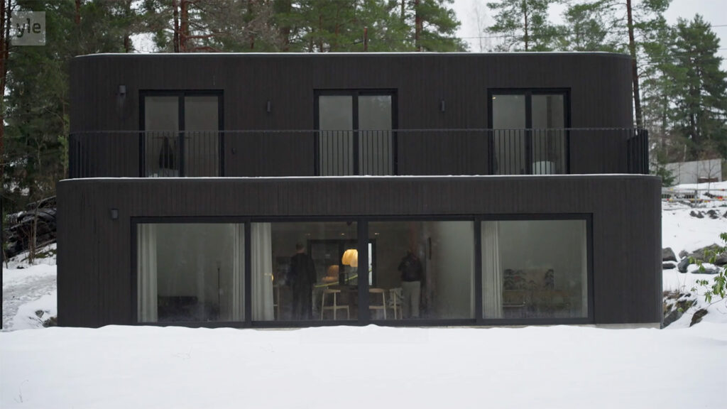 Stiligt - Ett modernt tvåvåningshus med stora fönster och mörk exteriör, omgiven av snö och träd, med människor synliga inuti på bottenvåningen.