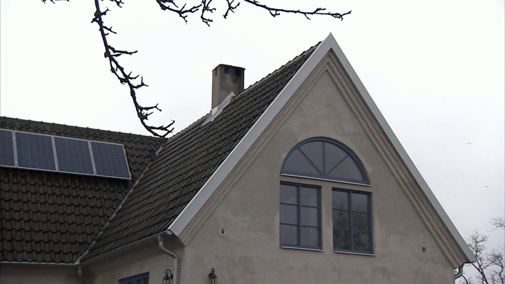 Stiligt - Ett beige hus med brant sadeltak, skorsten, välvt fönster, solpaneler på taket och kala trädgrenar i förgrunden.