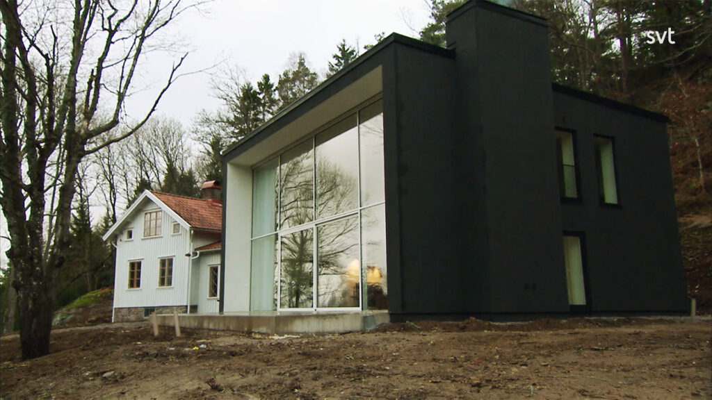 Stiligt - Ett modernt svart hus med stora fönster står bredvid ett äldre vitt trähus, omgivet av träd och barmark.