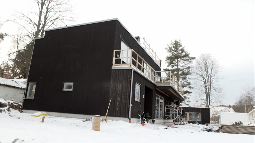 Stiligt - Ett modernt svart hus med platt tak under uppbyggnad på en snöig gård, med byggnadsställningar, verktyg och byggmaterial synliga.