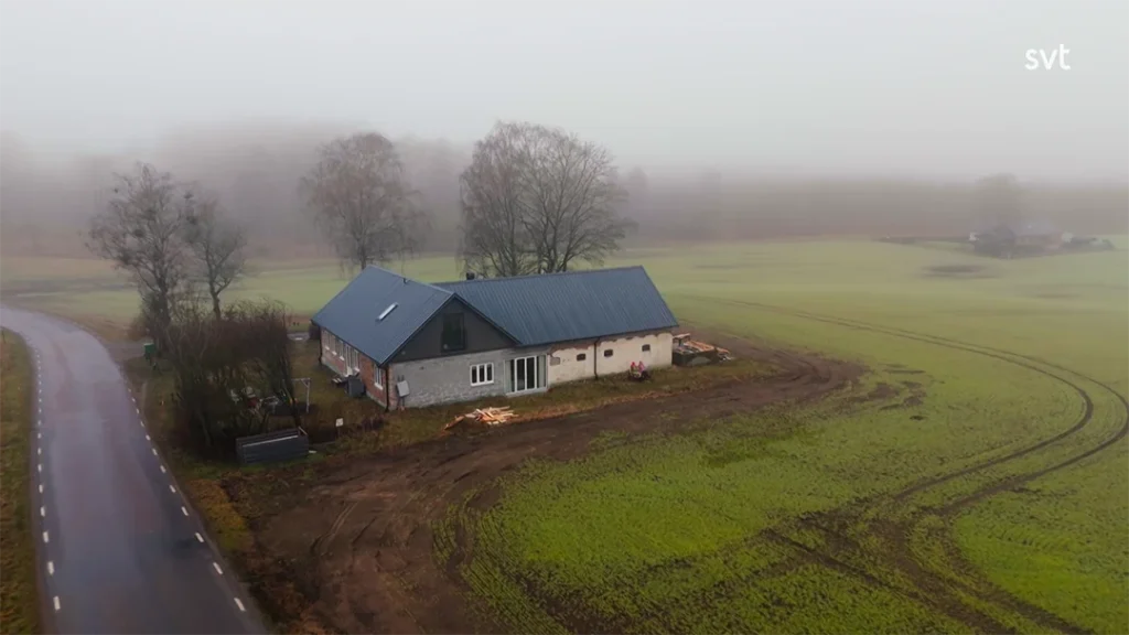 Stiligt - Ett hus med mörkt tak står bredvid en väg i Kverrestads dimmiga bygdelandskap, omgivet av åkrar och träd.