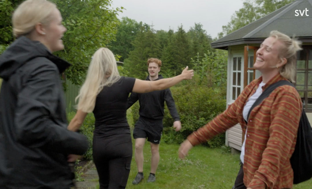 Stiligt - Fyra personer hälsar på varandra utomhus nära en liten byggnad vid Grönalund; två kvinnor ler, en person har sina armar utsträckta och träd i bakgrunden.